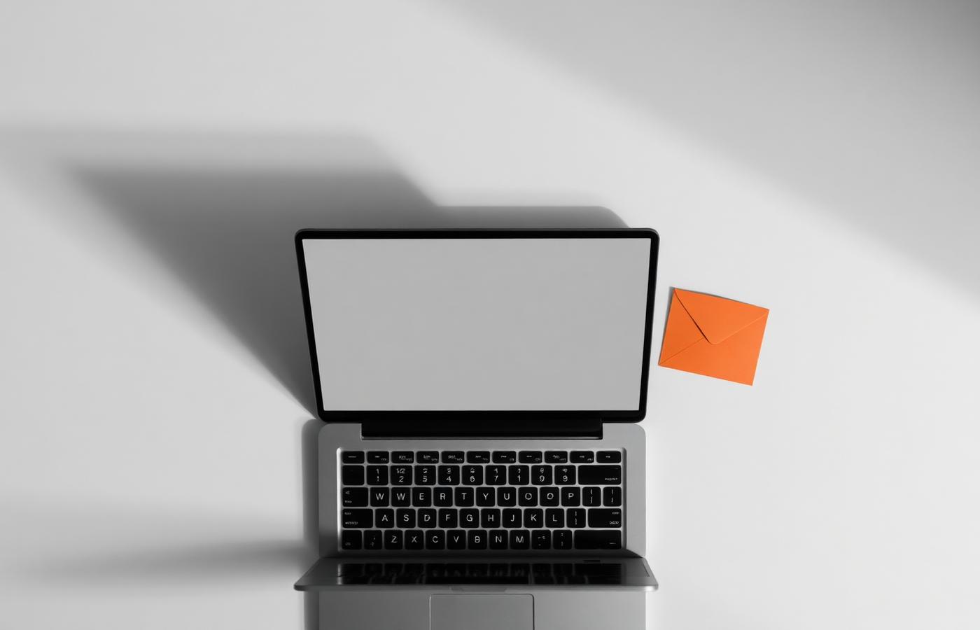 Top-down view of an open laptop with a blank screen beside a bright orange paper envelope on a light desk